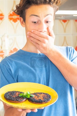 Photo for Young guy prepare alone lunch at home burning hamburgers and looking them with surprised expression. Concept of teenager independent. Home kitchen cooking concept people. Handsome teenager and meat - Royalty Free Image