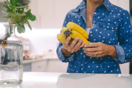 Photo for Close up of woman in the kitchen holding bananas  and living real life lifestyle. Healthy nutrition food and female. Dieting. - Royalty Free Image