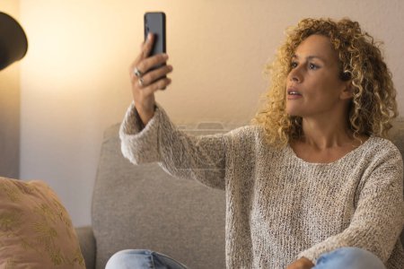 Photo for Woman busy in video call with mobile phone comfortably sitting at home on sofa. Indoor technology leisure activity. People using mobile phone application to take selfie picture for social media - Royalty Free Image