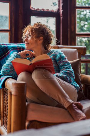 Photo for Alone  woman at home spending time reading a book and looking outside. Indoor leisure activity on holiday chalet vacation booking. Female people and knowledge education. Relaxation moment - Royalty Free Image