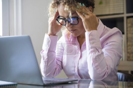 Photo for Painful headache on adult woman at work touching her temple. Thoughtful modern online worker female people in front a laptop at the desk. Business problems and lady worried. Overwork unhappy freelance - Royalty Free Image