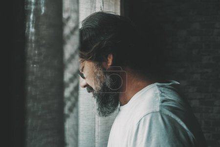 Photo for One lonely and depressed man with eyes closed stand up against the window at home. People and stress or desperation. Mature male with beard, sad alone indoor. Concept of unhappy person lifestyle. Mental - Royalty Free Image