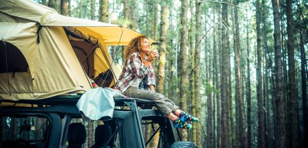 Photo for Freedom feeling and adventure lifestyle people. Traveler adult female chilling outside her car vehicle roof tent and enjoy nature sounds smiling. High trees forest green woods background. Scenic place - Royalty Free Image