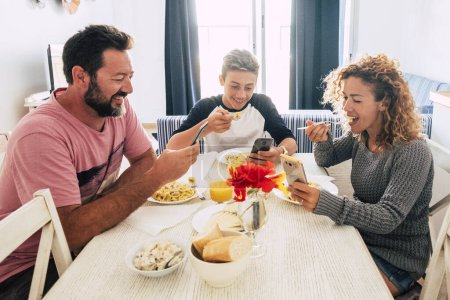 Téléchargez les photos : Joyeux jeune famille profiter du déjeuner ensemble à la maison en utilisant le téléphone mobile. Accro aux médias sociaux en ligne téléphone cellulaire les gens qui mangent dans le salon et regarder l'affichage s'amuser. Concept de smartphone addiction - en image libre de droit