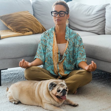 Foto de Mujer relajándose en casa con yoga rutinario posición de loto en la sala de estar con su mejor amigo perro tendido cerca de ella. Concepto de personas de estilo de vida mental fácil y saludable. Dueño del perro. Bienestar señora - Imagen libre de derechos