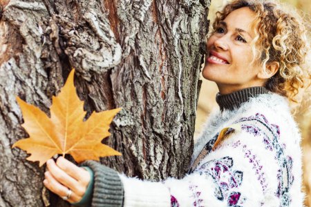 Photo for Close up portrait of serene expression woman hugging a tree and holding a yellow leaf enjoying outdoor leisure activity alone in autumn season. People and love for nature. Environment concept trees - Royalty Free Image