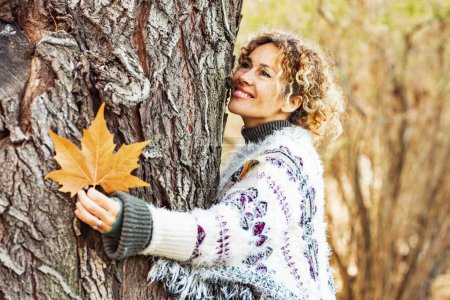 Téléchargez les photos : Femme adulte heureuse étreindre un arbre et sourire en automne activité de loisirs de plein air. Style de vie naturel et amour nature concept personnes. Bois de couleur jaune et des arbres en arrière-plan. Femme appréciant le parc - en image libre de droit