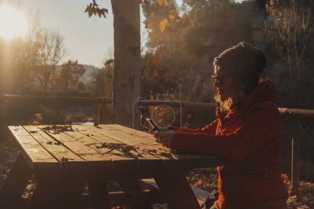 Téléchargez les photos : Femme avec veste rouge utilisant téléphone portable et connexion wifi extérieur assis sur un banc et une table en bois dans le parc lors d'un coucher de soleil incroyable automne doré. Femmes utilisant un appareil cellulaire loisirs - en image libre de droit