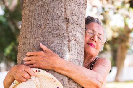 Téléchargez les photos : Vieille femme heureuse et sereine serrant un arbre dans le parc dans une activité de loisirs de plein air. Durabilité et environnement nature style de vie femmes. L'amour pour les arbres. Changement climatique et bien-être soucieux - en image libre de droit