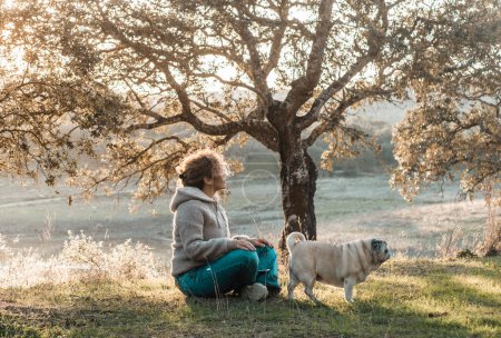 Téléchargez les photos : Femme assise sur l'herbe profitant d'un endroit calme et serein et le bien-être avec son meilleur ami chien. Ensemble pour toujours les gens la nature animale. Profiter du temps libre au lac. Tourisme rural. - en image libre de droit