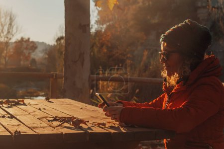 Téléchargez les photos : Femme utilisant un téléphone portable pour envoyer un message ou surfer sur le web dans le cadre d'une activité de loisirs de plein air seule assise sur un banc et une table en bois pendant le coucher de soleil doré en automne. Personnes et technologies de données extérieures - en image libre de droit