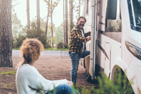 Photo for Adult couple enjoy outdoor leisure activity together outside a modern camper van motorhome. Travel people vacation lifestyle. Parking in a middle of nature with forest and trees in background. Freedom - Royalty Free Image