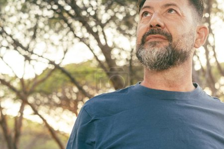 Photo for Close up portrait of confident adult male looking up. Bearded man with blue t-shirt and trees in background. People and outdoor leisure activity concept lifestyle. Serene expression on face. Copyspace - Royalty Free Image