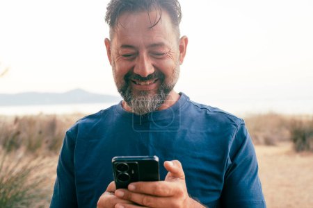 Photo for Front view of man using mobile phone with cheerful smile expression on face. Modern traveler people with connection and communication technology. Texting and messaging outdoor cellphone - Royalty Free Image
