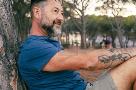 Téléchargez les photos : Homme adulte mature avec tatouage se détendre assis contre un coffre au parc et souriant regardant en face de lui. Personnes et activités de loisirs de plein air. Mode de vie sain. Voyages touristiques vacances - en image libre de droit