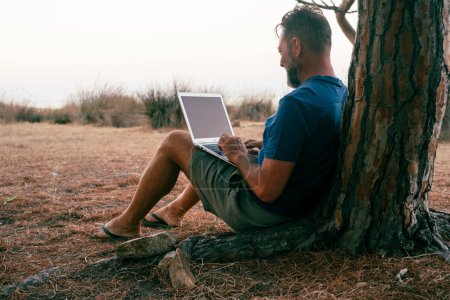 Photo for Digital nomad small business freedom lifestyle people concept outdoor office.  man entrepreneur freelance working on laptop sitting against a trunk at the park with nature around open space - Royalty Free Image