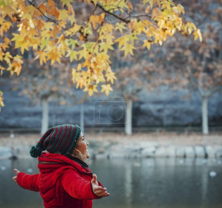 Foto de Vista lateral de mujer con chaqueta de invierno roja disfrutando de la actividad de ocio al aire libre en otoño con vista al lago. Gente turismo fin de semana concepto de viaje. Fondo de la naturaleza escénica. Libertad y estilo de vida. - Imagen libre de derechos