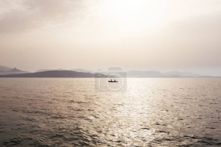 Téléchargez les photos : Misty humeur paysage marin avec bateau de pêcheurs au milieu de la mer et le littoral en arrière-plan sous la lumière du soleil de brouillard. - en image libre de droit