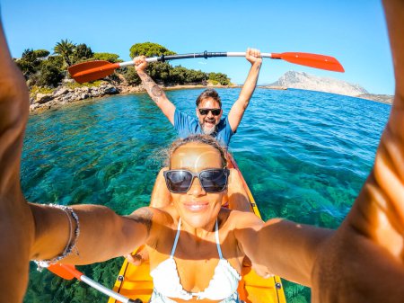 Téléchargez les photos : Couple heureux de caucasien adulte touriste homme et femme prenant des photos selfie à l'intérieur d'un canot de kayak avec de l'eau de mer bleu propre et le littoral en arrière-plan. Voyages et vacances d'été - en image libre de droit