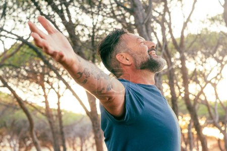Photo for Man outstretching arms in front of a high trees forest and sunset sunlight. Concept of environment and healthy lifestyle. People and love for nature. Male opening arms in front of sun. Travel day - Royalty Free Image