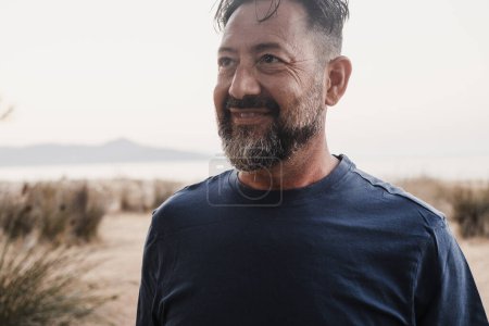 Photo for Side portrait of mature adult man with beard outdoor smiling and looking. Outdoors leisure activity. Casual male people with t-shirt and beach island scenic background destination. Vacation travel - Royalty Free Image