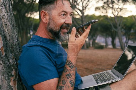Photo for Happy man do voice phone call outdoor at the park with computer on his legs sitting on the ground. Communication and outdoor connection. Travel digital nomad and open air office concept lifestyle - Royalty Free Image