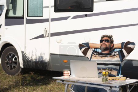 man digital nomad lifestyle working on alternative outside workplace desk with a camper van motor home house in background. People and office freedom. Modern small business concept job traveler