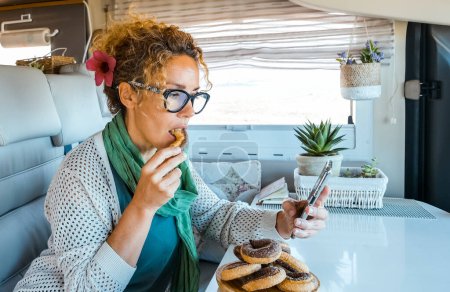 Photo for Adult woman sitting inside a camper van enjoy indoor leisure activity eating donuts sugar cakes and using mobile phone connection to surf the map and choose next trip destination. Vanlife holiday - Royalty Free Image
