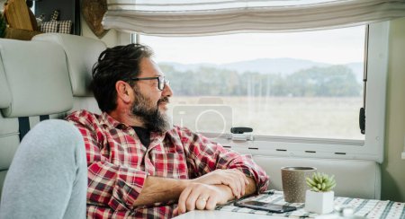 Photo for Adult man relaxing inside a camper modern motorhome looking outside the window enjoying the wild nature view. Alternative vacation and tiny house. Lifestyle vanlife. Modern traveler people - Royalty Free Image