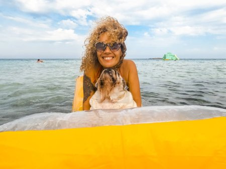 Téléchargez les photos : Femme heureuse s'amuser en vacances jouer et se détendre sur une lilo gonflable jaune avec son chiot de chien dessus et regardant la caméra. Les gens et le mode de vie animal. Vacances avec chien et loisirs en plein air - en image libre de droit