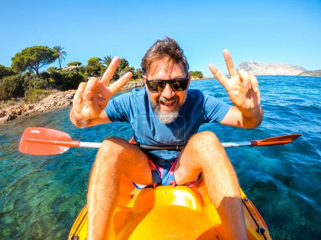 Téléchargez les photos : Homme joyeux s'amuser et poser pour une photo folle assis à l'intérieur d'un canot de kayak jaune avec de l'eau de mer et la côte en arrière-plan. Heureux touristes vacances d'été style de vie personnes faisant le tour - en image libre de droit