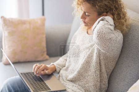 Téléchargez les photos : Femme assise à la maison sur le canapé en utilisant ordinateur portable fatigué et détendu. Ordinateur sans fil travailleurs. Femme adulte surfant sur le web seule à l'intérieur. Toucher le cou pour la douleur et la mauvaise posture malsain - en image libre de droit