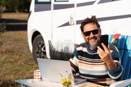 Happy traveler work on laptop computer outside a camper van and use phone call to connect and call. One man smile and enjoy cellphone calling witting outside at motor home alone at the table. 