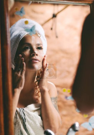 Photo for Attractive adult woman taking care of her skin face using cream product treatment in the bathroom looking in the mirror. Beauty care evening night routine female people at home. Spa wellbeing - Royalty Free Image