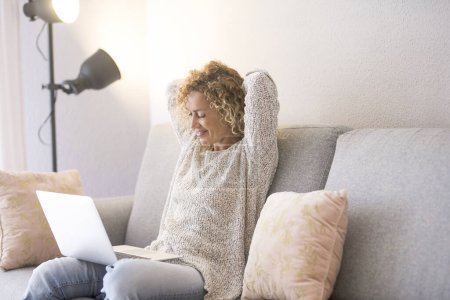 Téléchargez les photos : Femme utilisant un ordinateur portable assis sur le canapé dans la maison lumineuse. Les gens et la technologie en ligne. Femme moderne regardant l'ordinateur et détendre son dos et les épaules levant les bras sous la tête - en image libre de droit