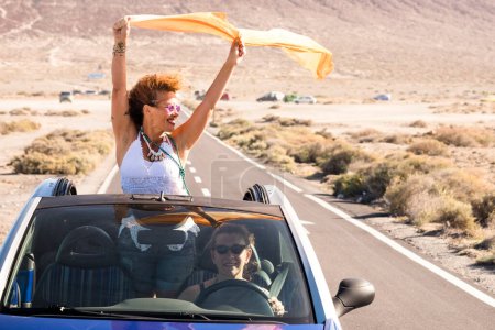 Photo for Travel and friendship concept lifestyle. Adult woman overjoyed staying outside the roof of a convertible car while her friend drive smiling. Concept of summer holiday vacation and road trip - Royalty Free Image