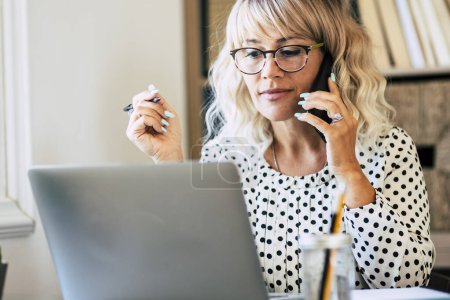 Téléchargez les photos : Femme entrepreneur adulte travaillant au bureau avec ordinateur portable et téléphone portable connexion cellulaire - dame d'affaires professionnelle avec des lunettes occupé à travailler intelligemment à la maison - emploi en ligne personnes modernes - en image libre de droit