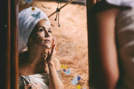 Foto de Mujer atractiva adulto utilizar crema de belleza en su cara mirando en el espejo en casa. Cuidado de la piel tratamiento anti envejecimiento en el baño. Mujeres con toalla blanca en la cabeza después del lavado - Imagen libre de derechos