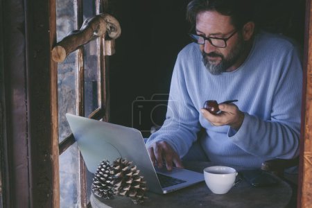 Téléchargez les photos : Homme mûr assis à la maison travaillant sur ordinateur portable et pipe à fumer. Alternative célibataire style de vie des hommes à l'intérieur dans chalet en bois cabine confortable. Télétravail en ligne - en image libre de droit
