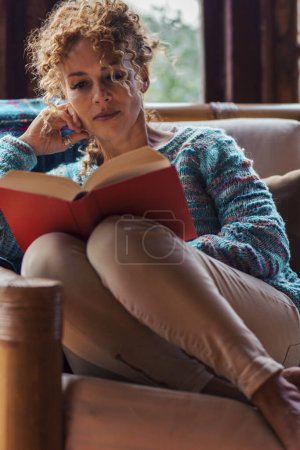 Photo for Relaxed woman alone at home sitting on a chair and reading a red book - female people indoor leisure relax activity read and enjoy time with windows and outdoors in background - Royalty Free Image