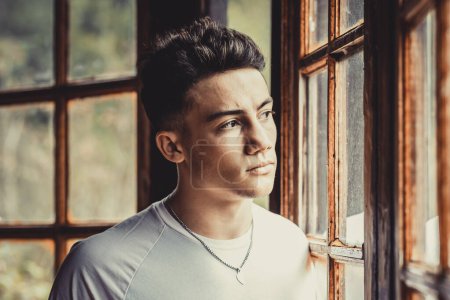 Photo for Handsome young man teenager looking serious outside the window at home. Teen problems concept lifestyle with worried boy reflected on the glass - Royalty Free Image
