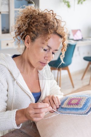 Photo for Adult caucasian woman at home doing crochet hobby activity at home decorating a shabby chic white pillow. Living room interior in background. Female people creating woolen decorations - Royalty Free Image