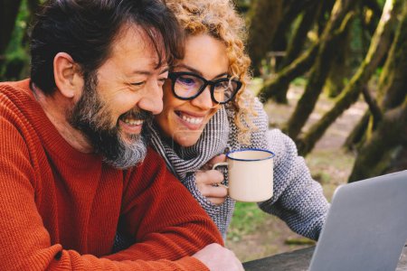Foto de Happy modern couple use together laptop computer outdoors with trees and woods in background - video call and smart remote working activity lifestyle concept with cheerful people and technology - Imagen libre de derechos