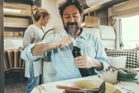 Photo for Adult traveler couple enjoy lunchtime inside a camper van. Nomadic alternative lifestyle. Travel people in summer holiday vacation ready to eat. Man opening wine and woman cooking - Royalty Free Image