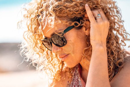 Photo for Middle age beautiful woman with curly hair and sunglasses in outdoor side portrait. Modern cute female people with tattoo relax at the beach under the summer sun. Healthy blonde people life - Royalty Free Image