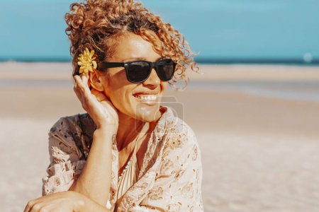 Photo for Portrait of happy tourist sitting and smiling at the beach with blue sky and ocean in background. Travel and tourism in summer holiday vacation. Female people with sunglasses on the sand - Royalty Free Image