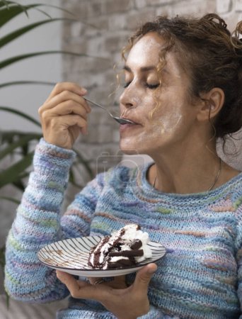Téléchargez les photos : Femme à la maison appréciant manger un gâteau savoureux avec de la crème et du chocolat avec expression de plaisir sur le visage. Les femmes se détendent à la maison. Concept de nourriture et de bonheur. Jeune femme adulte sur le canapé - en image libre de droit