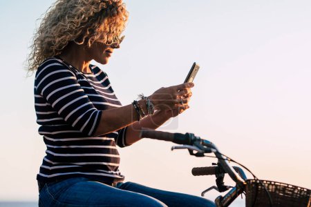 Foto de Vista lateral de la mujer adulta usando la conexión del teléfono móvil a la mensajería sentada en una bicicleta clásica. Medio ambiente de transporte verde estilo de vida. Las mujeres disfrutan de ocio al aire libre y conexión a Internet - Imagen libre de derechos