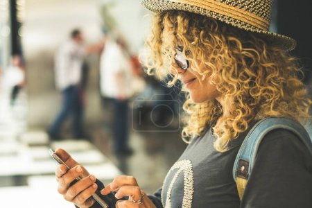 Photo for Side portrait of pretty  woman using mobile phone connection to send messages and chat. Traveler concept people with backpack and hat. Blonde long curly hair female use cellular at airport - Royalty Free Image