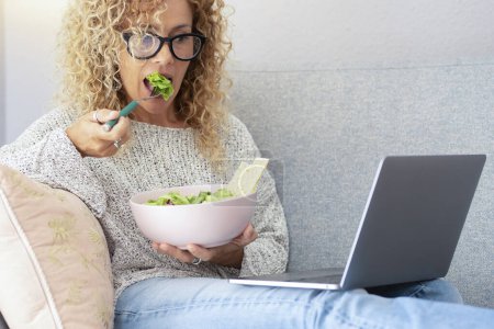 Photo for Healthy lifestyle and modern online work activity concept. A woman eating salad and looking laptop sitting on the sofa. One female people using computer alone at home. Indoor leisure web activity - Royalty Free Image